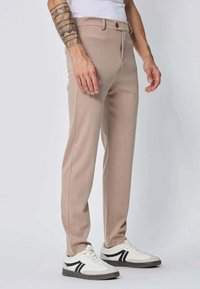 Pantalons beige ajustés avec une texture lisse et des lignes épurées. Présente un bouton à l'avant et des poches, associés à des baskets blanches avec des accents noirs.