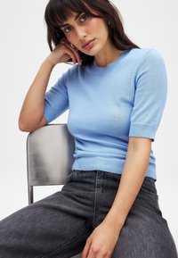 Svetlo modra kratkolužečni pulover iz mehkega materiala z okroglim izrezom in rebrastim robom, v kombinaciji s temno sivo hlačami z visokim pasom.