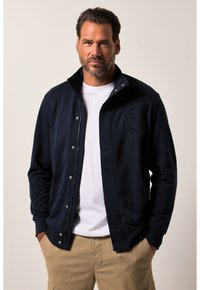 Giacca blu navy con cerniera e bottoni a pressione, caratterizzata da una toppa logo testurizzata sul petto, abbinata a una t-shirt bianca sotto.