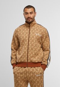 Brown patterned zip-up jacket with high collar, matching pants, and navy side stripes. Features logo detail and textured fabric.