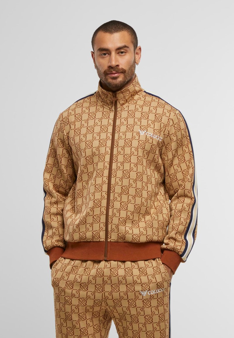 Brown patterned zip-up jacket with high collar, matching pants, and navy side stripes. Features logo detail and textured fabric.