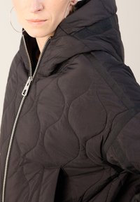 Veste matelassée noire avec fermeture éclair, capuche et tissu texturé au fini mat. Accentuée par des bretelles noires élégantes sur les épaules.