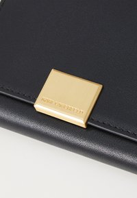 Crna kožna novčanica s zlatnim pravokutnim okovom utisnutim s natpisom "KARL LAGERFELD" blizu ruba. Glatka tekstura i minimalistički dizajn.