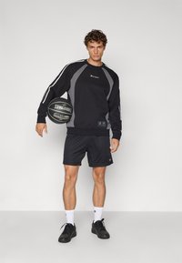 Sudadera negra con detalles en gris, que presenta un logotipo y puños acanalados, combinada con pantalones cortos de malla negros y zapatillas negras, sosteniendo un baloncesto.