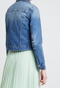 Osoba oblečená v modré denimové bundě přes světle zelenou plisovanou sukni, stojící před jednoduchým bílým pozadím.