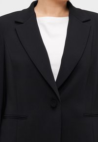 Czarna marynarka z kołnierzem z klapami, zapinana na pojedynczy guzik, z dwoma przednimi kieszeniami z patkami i gładką fakturą. Nosi się ją na białą bluzkę.