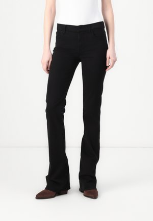 Person iført sorte bootcut bukser og brune spidse sko, der står mod en hvid baggrund.
