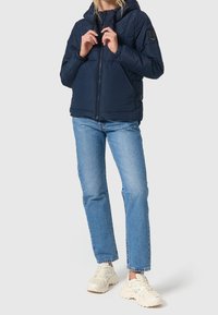 Marineblå pufferjakke med hætte, der har en frontlynlås, elastiske manchetter og en tekstureret overflade. Båret med lyseblå jeans og sneakers.