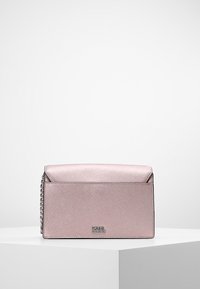 Vaaleanpunainen, teksturoitu synteettinen käsilaukku kääntyvälla yläosalla ja ketjusangalla. Edessä metallinen logo ja sileä pinta.