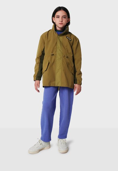 Olivgrüne, wasserdichte Jacke mit Reißverschluss, Kordelzug in der Taille und zwei Fronttaschen. Kombiniert mit blauen Hosen und weißen Sneakers.