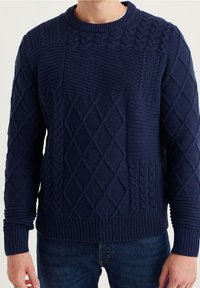 Man som bär en marinblå kabelstickad tröja med diamant- och flätmönster, tillsammans med mörkblå jeans, mot en enfärgad bakgrund.