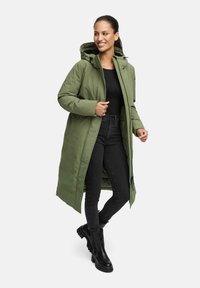 Lange groene gewatteerde jas met een capuchon, voorzien van een ritsluiting, zijzakken en een rechte snit, gecombineerd met zwarte jeans en laarzen.