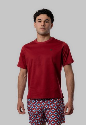 Giovane uomo in piedi, indossa una maglietta rossa a maniche corte semplice e pantaloncini geometrici a motivi rossi, blu e bianchi con sfondo grigio.