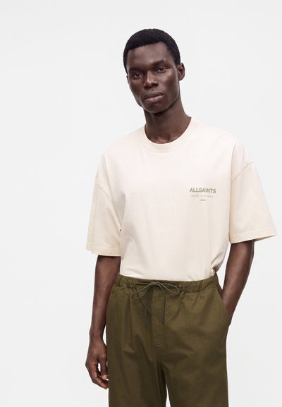Νέος άνδρας που φοράει μπεζ t-shirt AllSaints και χακί παντελόνι με κορδόνι, στέκεται με το ένα χέρι στην τσέπη πάνω σε απλό φόντο.