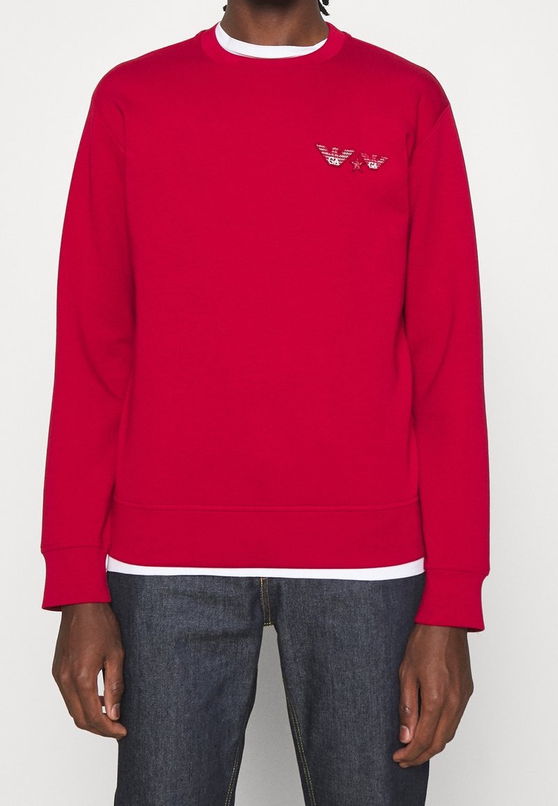 Personne portant un sweat rouge à manches longues avec un logo brodé sur la poitrine, associé à un jean bleu foncé et un sous-vêtement blanc visible au niveau du col et de l’ourlet.