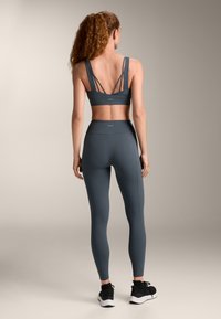 Szürke sportmelltartó széles, keresztbe kötött pántokkal és azonos anyagú, magas derékú leggings-szel, sima textúrával és testhezálló kialakítással. Fekete sportcipők.