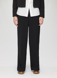 Pantalon large noir avec une texture lisse et un devant plissé, associé à une chemise blanche à boutons sous une veste noire cropped.