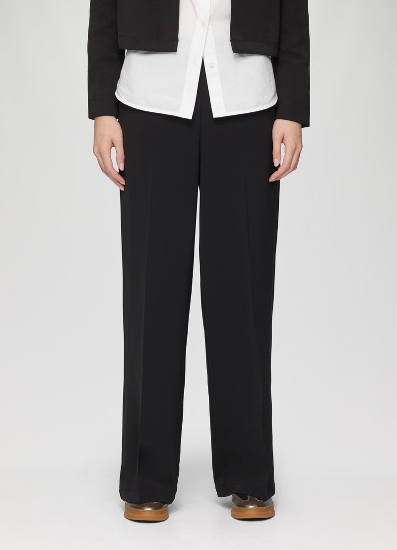 Pantalon large noir avec une texture lisse et un devant plissé, associé à une chemise blanche à boutons sous une veste noire cropped.