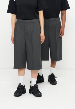 Twee mensen dragen zwarte T-shirts, grijze op maat gemaakte shorts tot net over de knie, witte sokken en zwarte sneakers, staand naast elkaar.