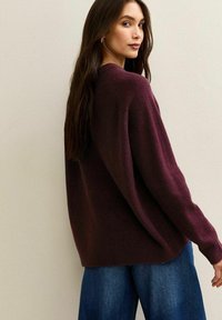 Maglione in lana bordeaux con una vestibilità comoda, maniche lunghe e polsini a coste; abbinato a jeans larghi blu, vista da dietro.