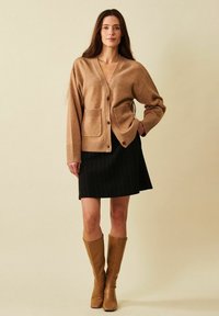 Nainen, joka pukeutunut vaaleanruskeaan neulejakkuun, mustaan hameeseen ja polven korkuisiin vaaleanruskeisiin saappaisiin, seisoo käsi taskussa yksiväristä taustaa vasten.