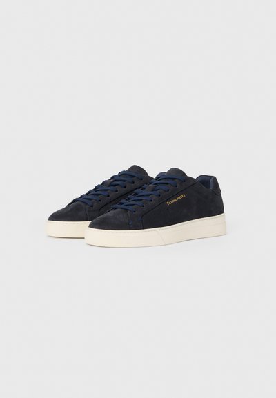 Tumši zilo zamšādas apavu pāris ar atbilstošām mežģīnēm, baltām gumijas zolēm un zelta "Filling Pieces" logotipu sānos.