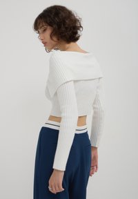 Femme aux cheveux bouclés courts regardant vers le bas, portant un pull court en coton blanc à côtes et un pantalon bleu marine devant un fond uni.