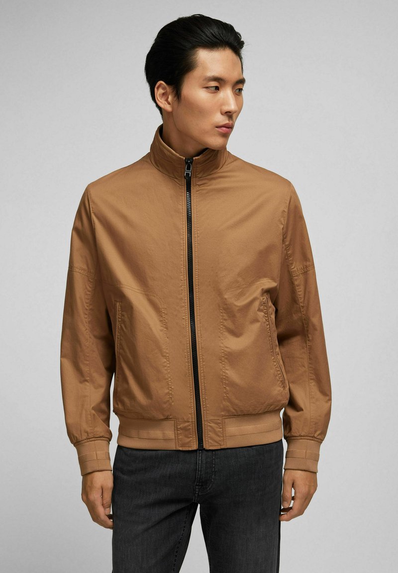 Tan Bomberjacke mit hohem Kragen und Reißverschluss. Verfügt über gerippte Abschlüsse und einen Saum, aus leichtem Stoff mit glatter Textur.