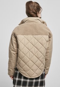 Gepolsterte beigefarbene Jacke mit einem weichen, kuscheligen braunen Kragen. Merkmale einen abgerundeten Saum und ein kariertes schwarz-weißes Muster darunter.
