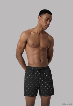Homme musclé portant un short de bain noir Calvin Klein, debout les mains derrière le dos, regardant vers le bas sur un fond gris uni.