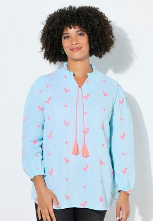 Kobieta o kręconych włosach, ubrana w jasnoniebieską bluzkę z różowymi wzorami i troczkami z frędzlami, stoi na białym tle.