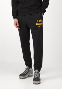 Sorte joggebukser med gult "Lakers"-logo og Nike-merke, med elastisk midje og innsnevrede mansjetter, brukt med grå treningssko.