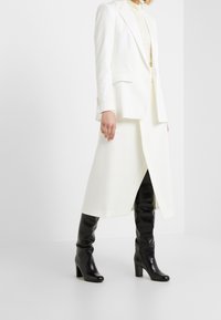 Blazer bianco su misura sopra una maglia crema, abbinato a una gonna midi bianca e stivali neri al ginocchio. Tessuti lisci e raffinati e silhouette strutturata.