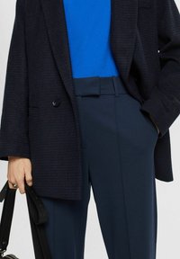 Person som bär en mörk rutig blazer, en ljusblå topp, marinblå byxor med bältesdetaljer, och håller en svart handväska med bred rem.