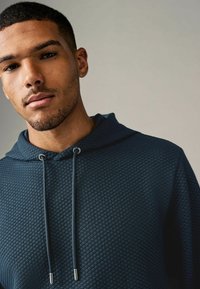Sudadera con textura en azul oscuro con un patrón de diamantes, que presenta una capucha con cordón y acentos metálicos en los extremos del cordón.
