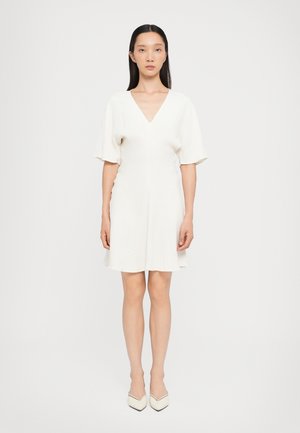 Vestido corto blanco con escote en V, mangas cortas, tela texturizada y falda acampanada. Combinado con tacones blancos puntiagudos.