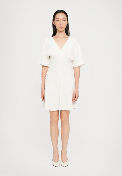 Vestido corto blanco con escote en V, mangas cortas, tela texturizada y falda acampanada. Combinado con tacones blancos puntiagudos.
