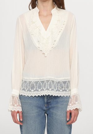 Blouse blanche transparente avec col en V volanté, détails en dentelle florale sur le col, les poignets et l'ourlet, portée avec un jean bleu.