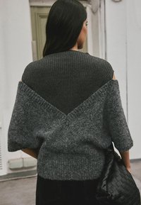 Grauer Strickpullover mit geripptem oberen Bereich, weiten Ärmeln und einem v-förmigen Rückendesign. Getragen mit einer strukturierten schwarzen Tasche.