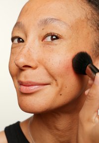 Pudderblushbørste påfører rosafarget blush på en jevn, fregnet kinn. Huden har en naturlig glød; bakgrunnen er enkel og lysfarget.