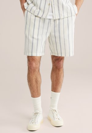 Hombre con pantalones cortos blancos texturizados con rayas verticales azules, combinados con calcetines blancos y zapatillas blancas, de pie con las manos en los bolsillos.