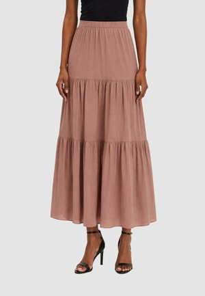 Donna che indossa una gonna lunga a balze color rosa polvere con fascia elastica in vita e sandali neri con cinturino alla caviglia e tacco alto.