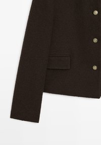 Donkerbruine, gestructureerde mouwen van een jas, met een voorste flapzak en gouden decoratieve knopen op een witte achtergrond.