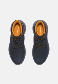 Granatowe zamszowe buty z żółtymi wkładkami, z okrągłymi sznurowadłami i teksturowaną gumową podeszwą. Akcentowane przeszycia podkreślają design.