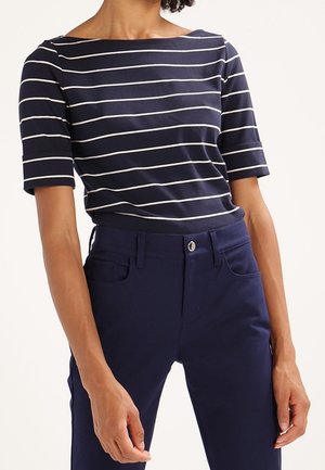 Person, die ein marineblau-weiß gestreiftes Hemd mit ellbogenlangen Ärmeln und marineblaue Hose vor einem einfarbigen Hintergrund trägt.
