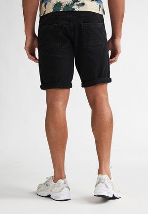 Shorts en jean noir avec revers roulés, deux poches arrière, associés à des baskets blanches. Le tissu semble robuste et légèrement texturé.