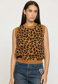 Femme aux longs cheveux noirs portant un haut en tricot sans manches à imprimé léopard brun et un jean bleu avec un patch sur la poche, se tenant devant un fond uni.