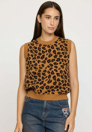 Femme aux longs cheveux noirs portant un haut en tricot sans manches à imprimé léopard brun et un jean bleu avec un patch sur la poche, se tenant devant un fond uni.