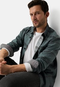 Blaugrünes kariertes Hemd mit grauen Ärmeln, getragen über einem weißen T-Shirt. Das Modell sitzt mit verschränkten Armen und zeigt den Lagenlook und die Knopfdetails.