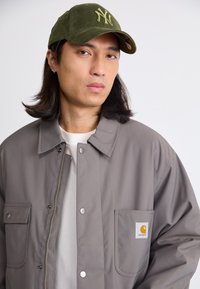 Grå zip-jakke med to frontlommer og et logo patch, parret med en olivengrøn kordfløjlskasket med broderede "NY" initialer.